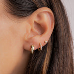 Close up of model wearing small gold huggie earrings featuring a diamond stripe in the center in her first piercing, and then a diamond huggie hoop on her second piercing and a tiny diamond stud in her third piercing