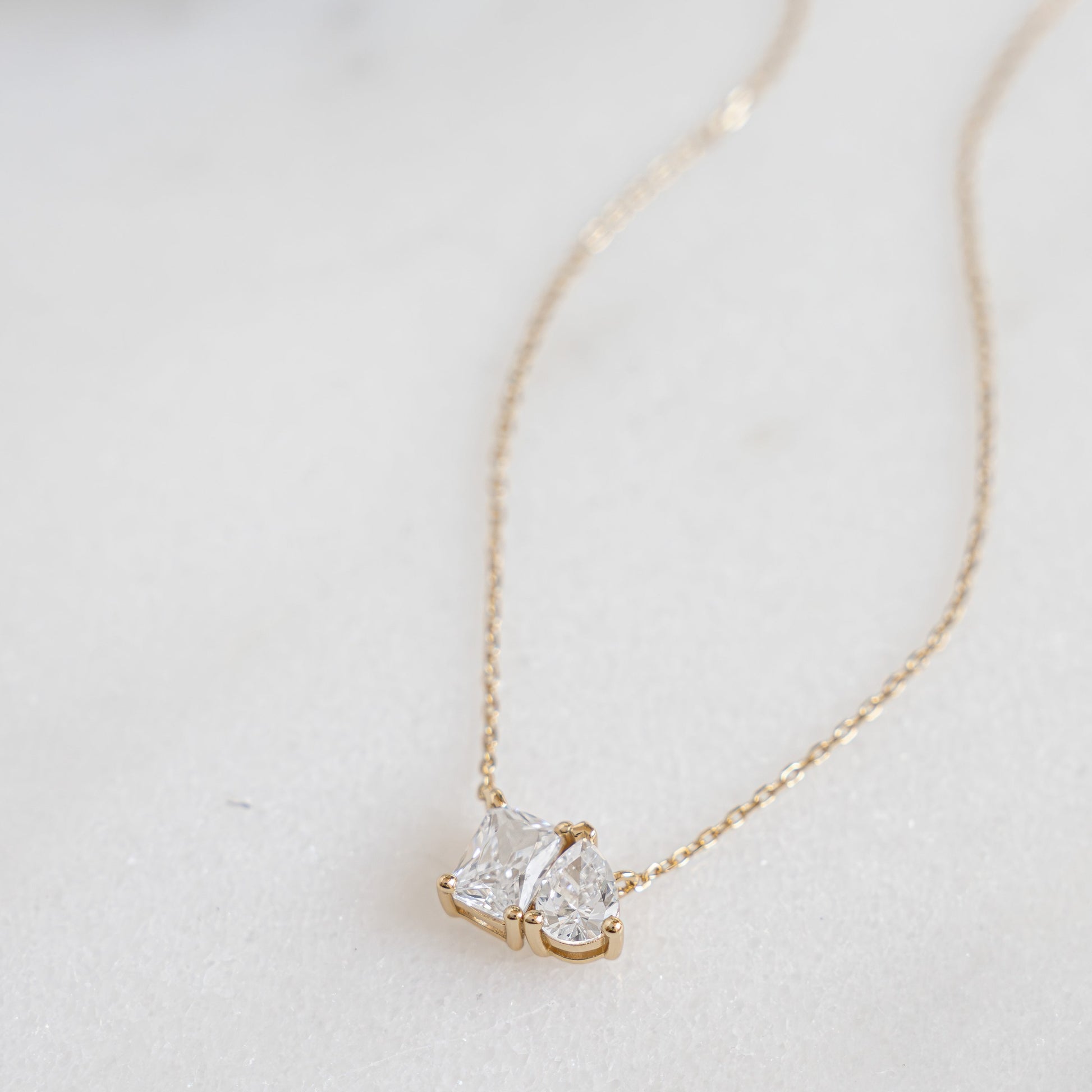 A delicate gold necklace featuring two gemstones, an emerald cut and a pear-shaped stone, laid flat.