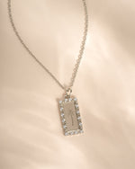 A silver-toned rectangular pendant necklace with a halo of cz stones and an engraved palm tree design, displayed on a soft beige fabric background.