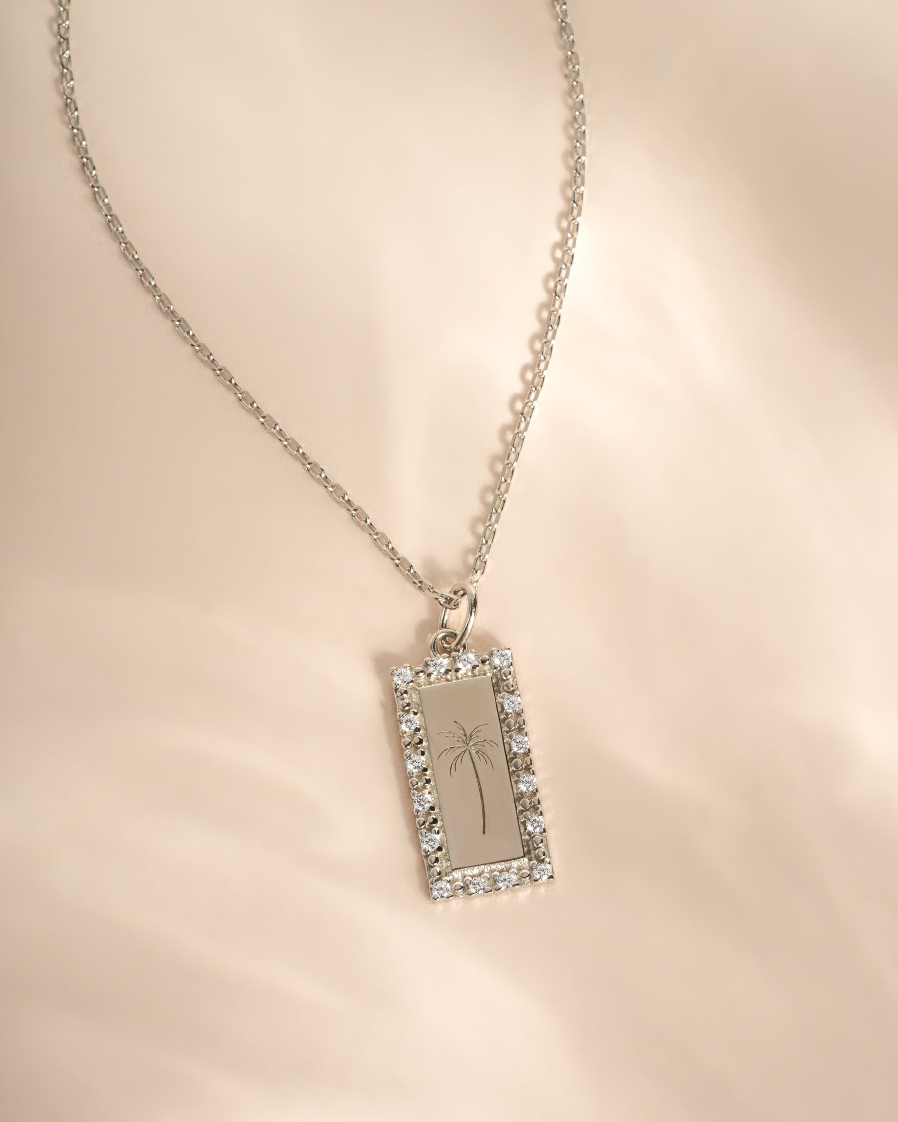 A silver-toned rectangular pendant necklace with a halo of cz stones and an engraved palm tree design, displayed on a soft beige fabric background.
