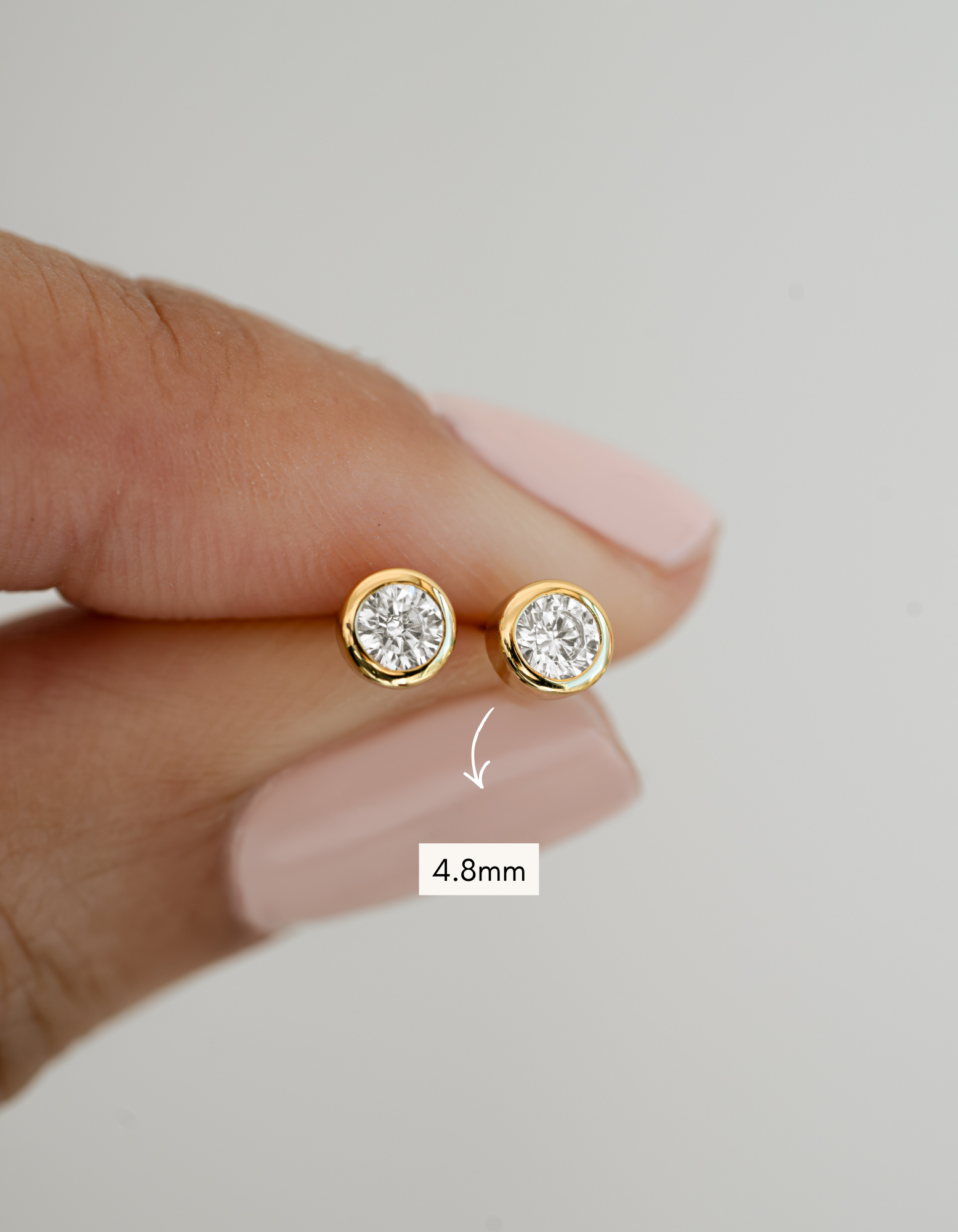 Gold stud earrings with diamond-like stones held between fingers, size indicated as 4.8mm.