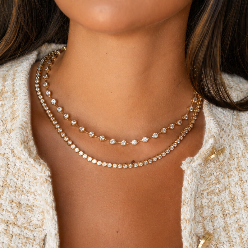 A close-up of a woman wearing a double-strand gold chain necklace with small clear stones, styled with a white ribbed tank top and a textured cream tweed jacket.
