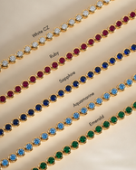 Five gold-tone tennis bracelets laid diagonally on a beige background, each set with different colored round stones and labeled with text: White CZ, Ruby, Sapphire, Aquamarine, and Emerald.