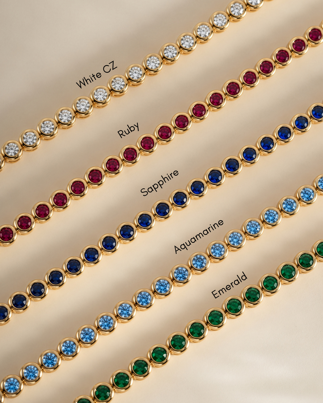 Five gold-tone tennis bracelets laid diagonally on a beige background, each set with different colored round stones and labeled with text: White CZ, Ruby, Sapphire, Aquamarine, and Emerald.