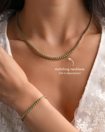 A close-up of a model wearing a delicate gold necklace with green stones and a matching bracelet on her wrist, styled with a white lace top. Text on the image indicates the matching necklace is in the description.