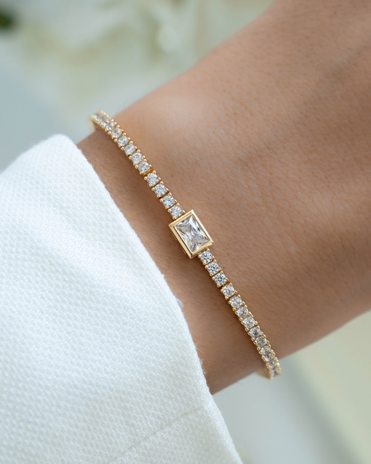 Gold Sami Jewels bezeled emerald-cut charm tennis bracelet worn on a wrist with delicate chain and gemstone accents.