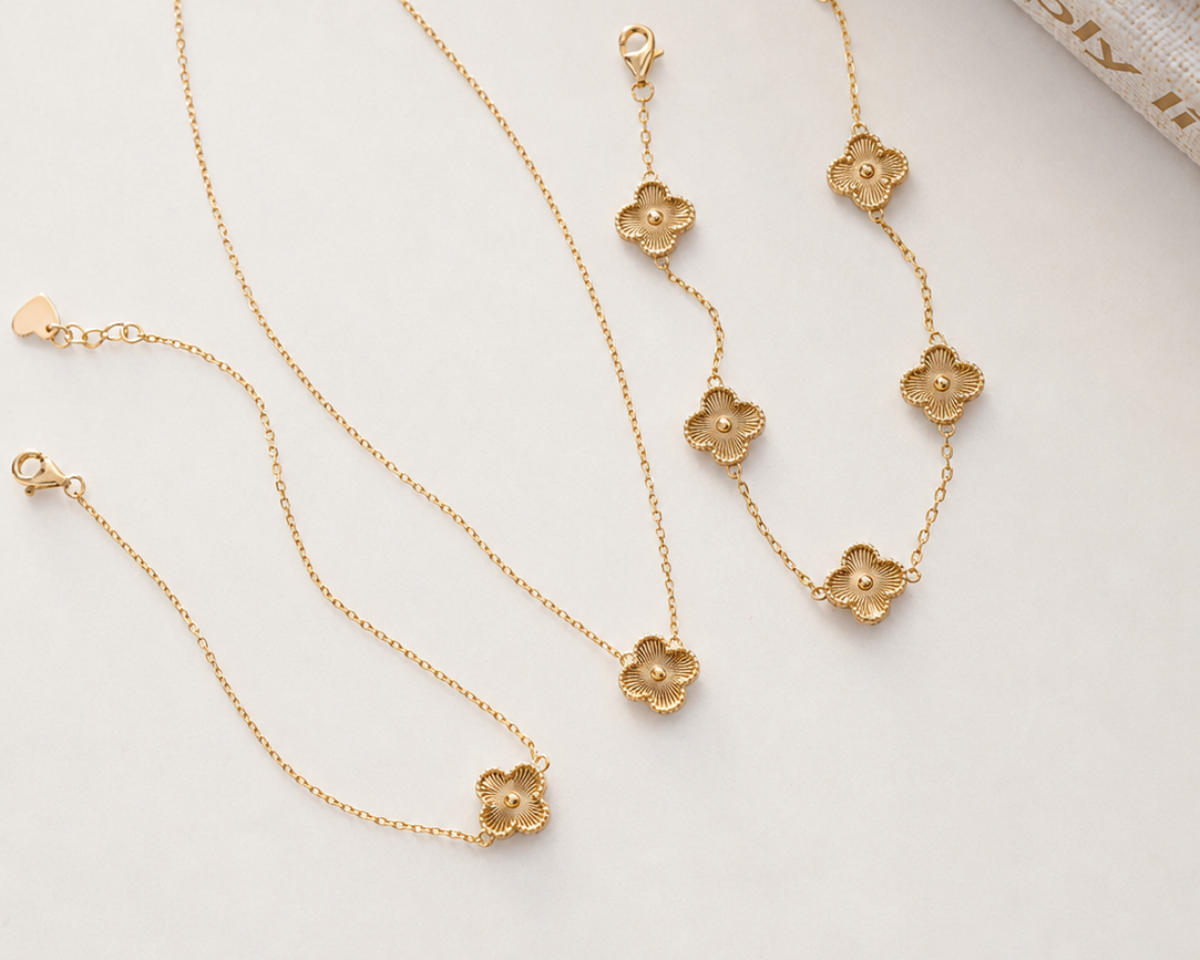 A gold jewelry set featuring a delicate chain necklace, bracelet, and anklet, each adorned with a small five-petal flower charm, arranged on a light beige textured surface.
