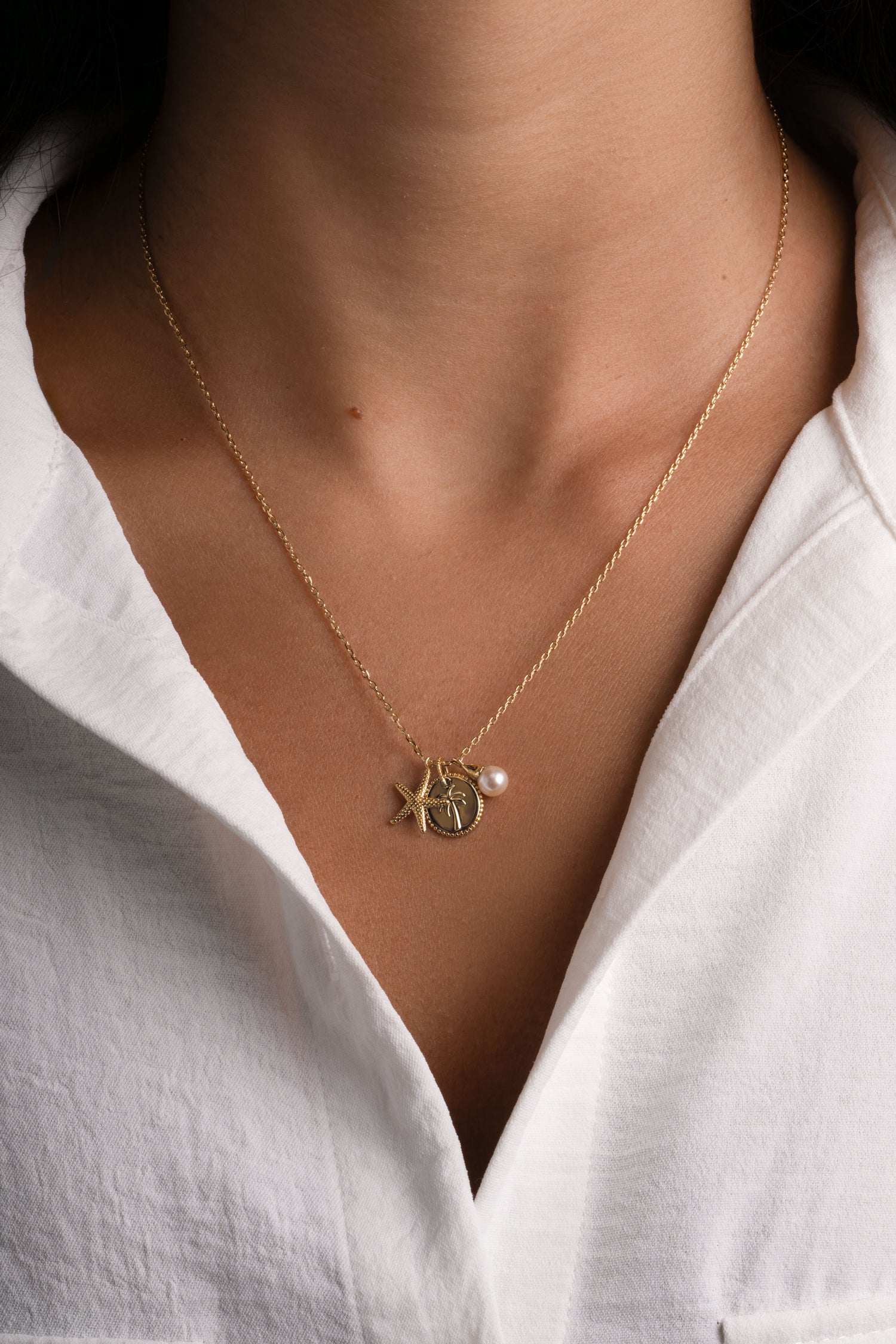 A close-up of a person wearing a delicate gold chain necklace with a starfish and palm tree pendant, along with a small pearl accent, styled with a white collared shirt.