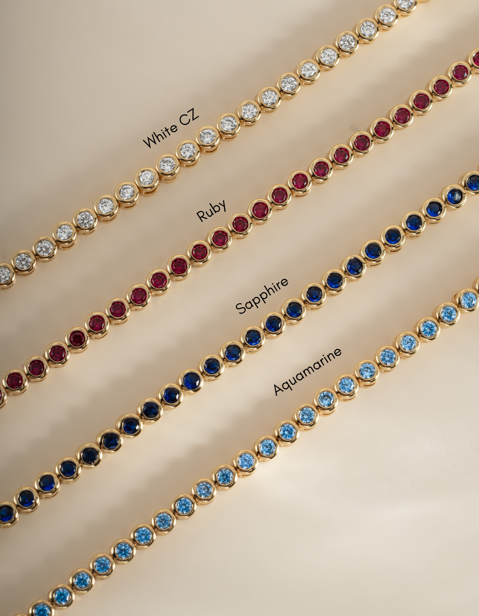 Four gold tennis bracelets laid diagonally on a beige background, each set with different gemstones and labeled 'White CZ', 'Ruby', 'Sapphire', and 'Aqua'.