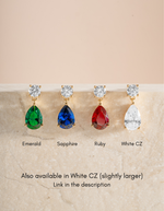 Four teardrop-shaped gemstone drop earrings displayed on a light stone surface against a soft beige background. From left to right, the labels read Emerald, Sapphire, Ruby, and White CZ.