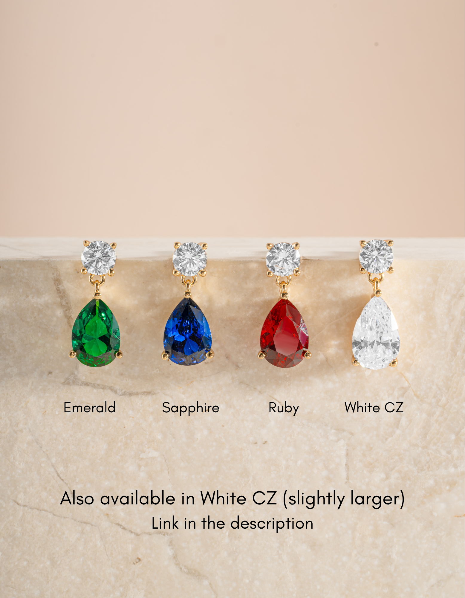 Four teardrop-shaped gemstone drop earrings displayed on a light stone surface against a soft beige background. From left to right, the labels read Emerald, Sapphire, Ruby, and White CZ.