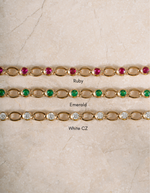 Three gold tennis-style bracelets laid flat on a light beige stone surface, labeled Ruby, Emerald, and White CZ. The top bracelet has pinkish-red stones, the middle has green stones, and the bottom has clear stones.