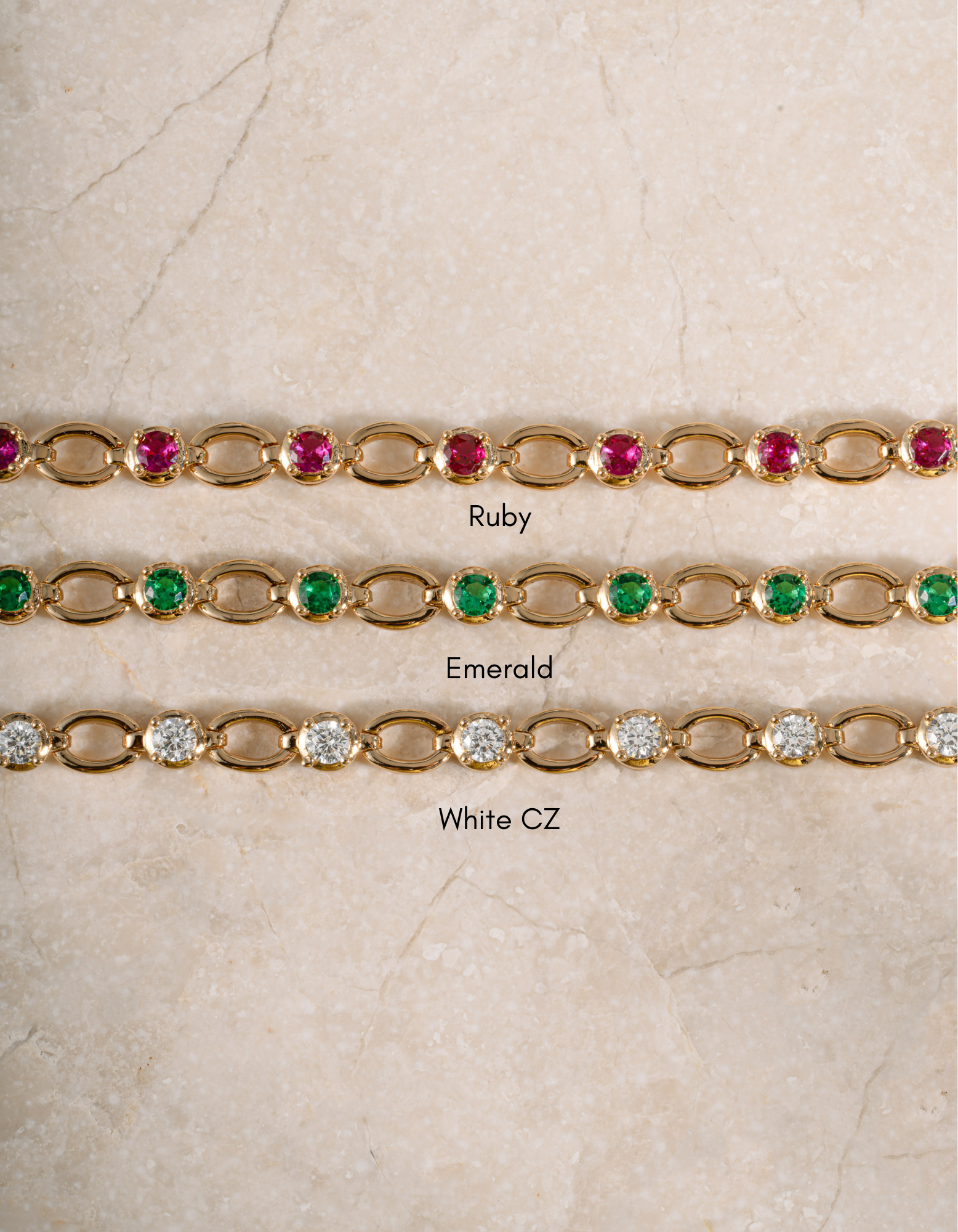 Three gold tennis-style bracelets laid flat on a light beige stone surface, labeled Ruby, Emerald, and White CZ. The top bracelet has pinkish-red stones, the middle has green stones, and the bottom has clear stones.