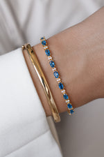 Two bracelets featuring a gold bangle and a tennis bracelet with oval sapphires and round diamond stones modeled on a wrist.