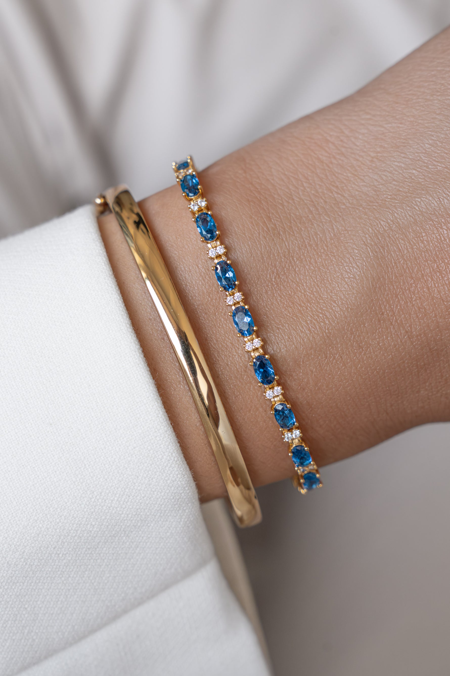 Two bracelets featuring a gold bangle and a tennis bracelet with oval sapphires and round diamond stones modeled on a wrist.