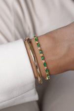 Two bracelets styled on a wrist featuring an oval-cut emerald tennis bracelet with alternating round white stones and a gold bangle.
