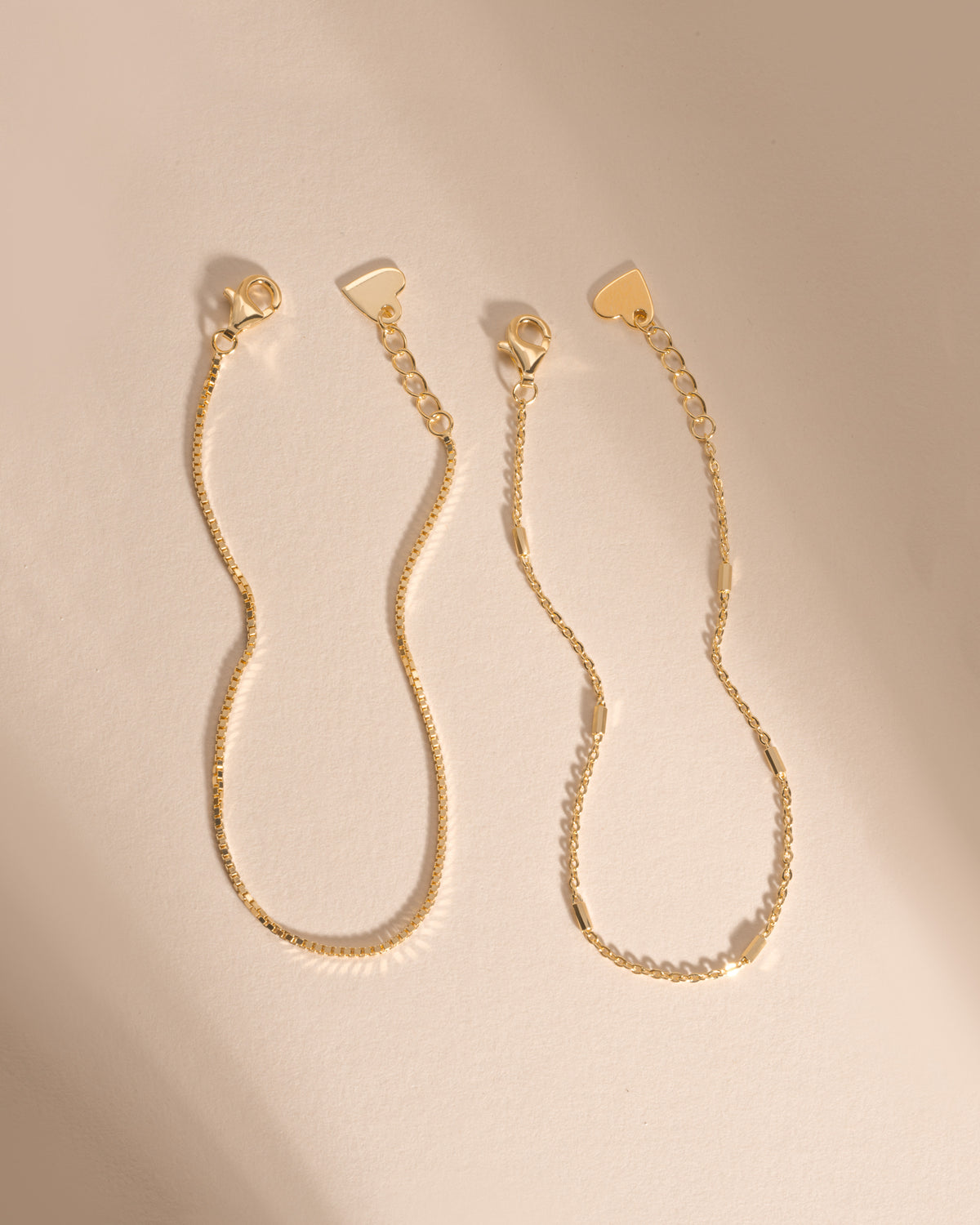 Two gold bracelets laid side by side, showing their lobster clasps, extenders and heart-shaped tags.