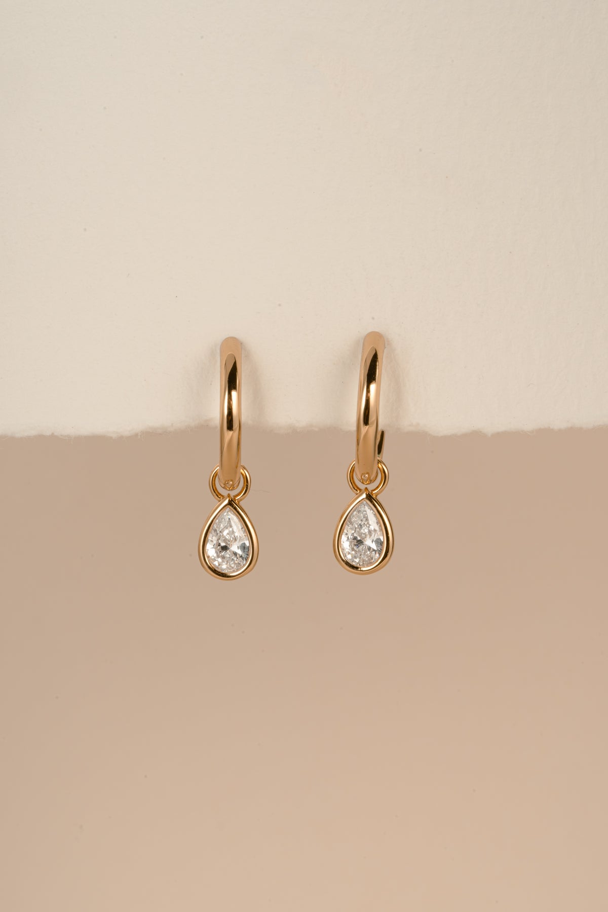 Pair of gold plated pear CZ charm earrings hanging against a warm neutral background.