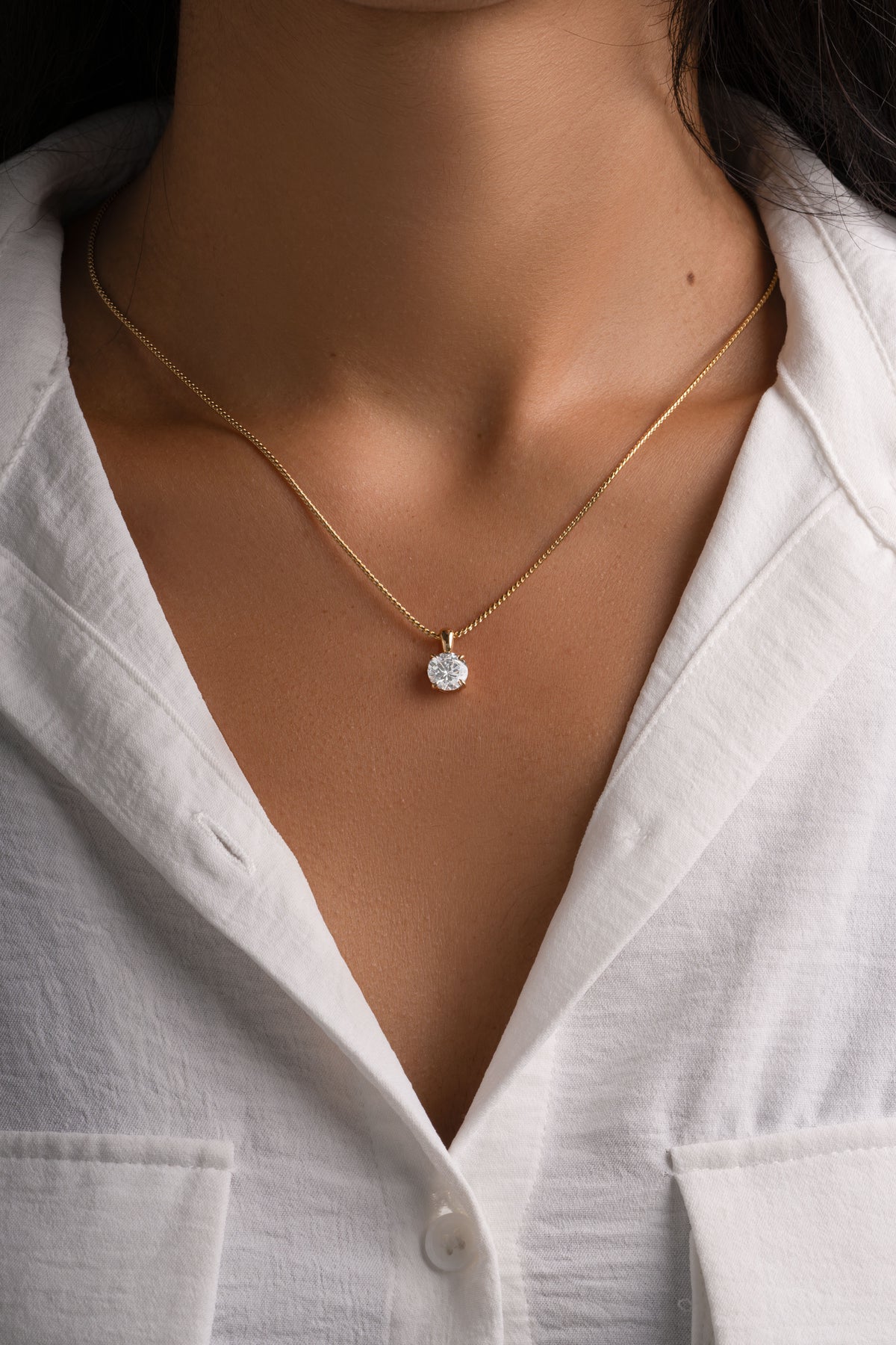 Gold round solitaire pendant worn alone on a model in a white shirt.