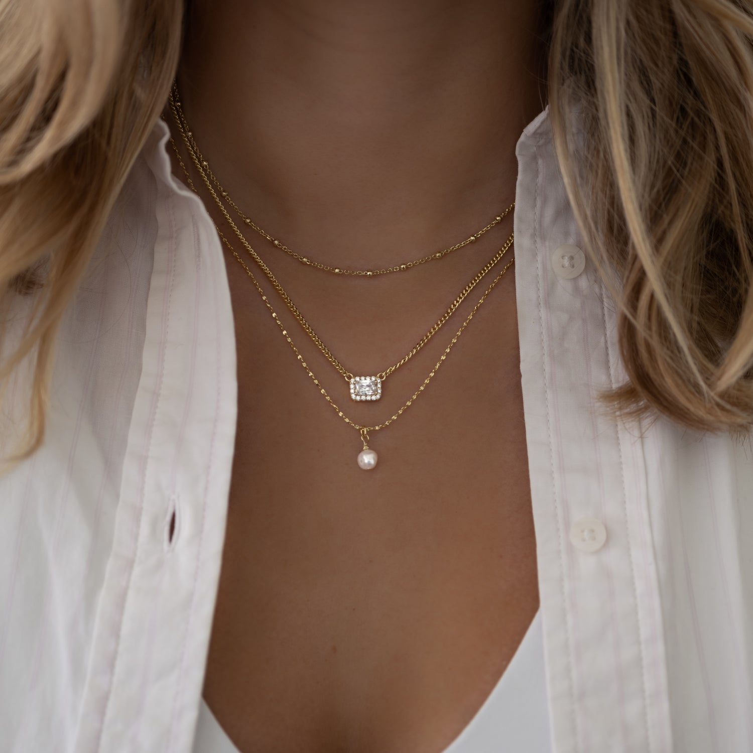 gold necklace stack featuring a freshwater pearl necklace, a halo emerald cut cuban link necklace, and a simple chain.
