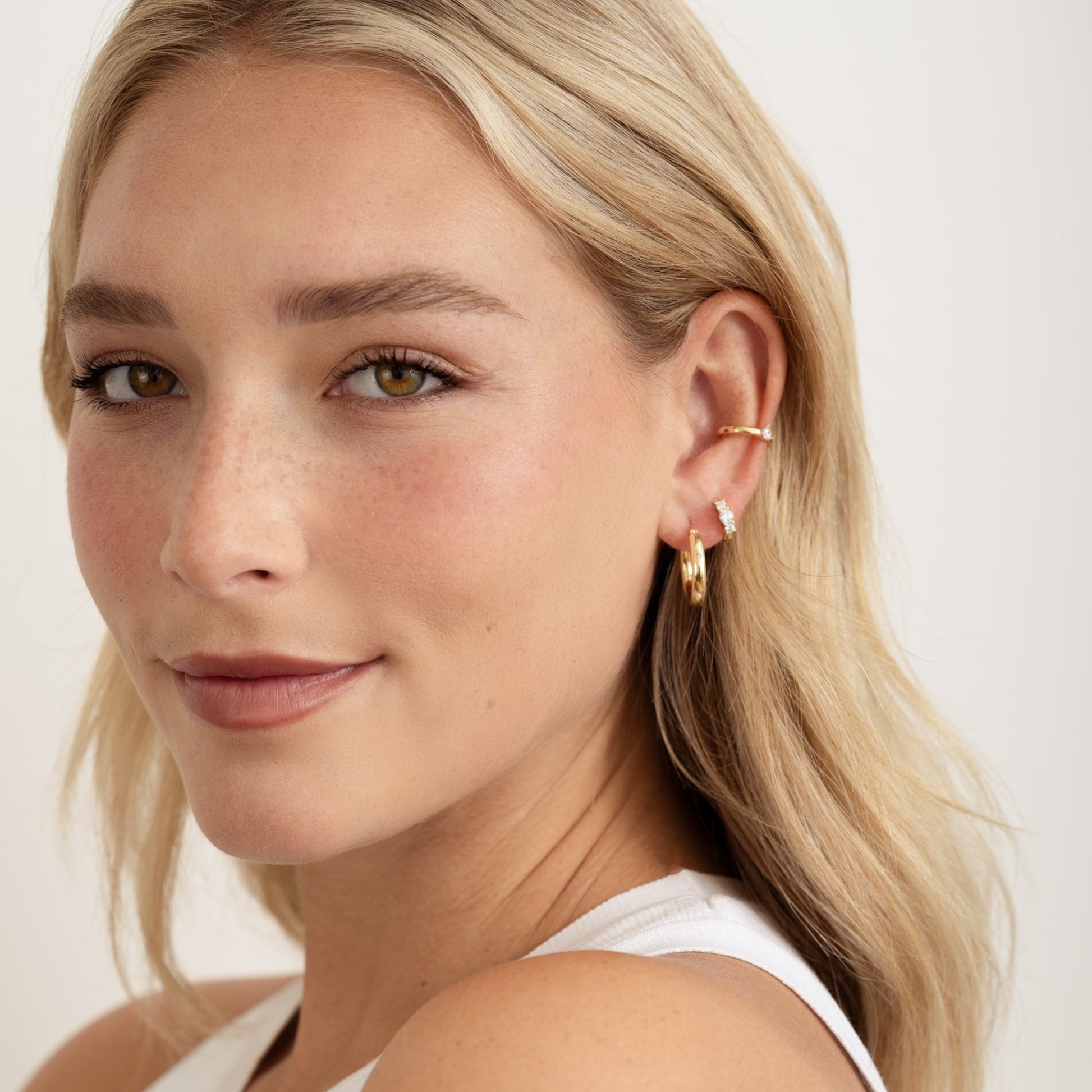 a model wearing gold hoop earrings, small huggies in her second piercing and a gold minimalist ear cuff