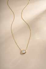 Gold east-west oval bezel pendant necklace arranged on a cream background.