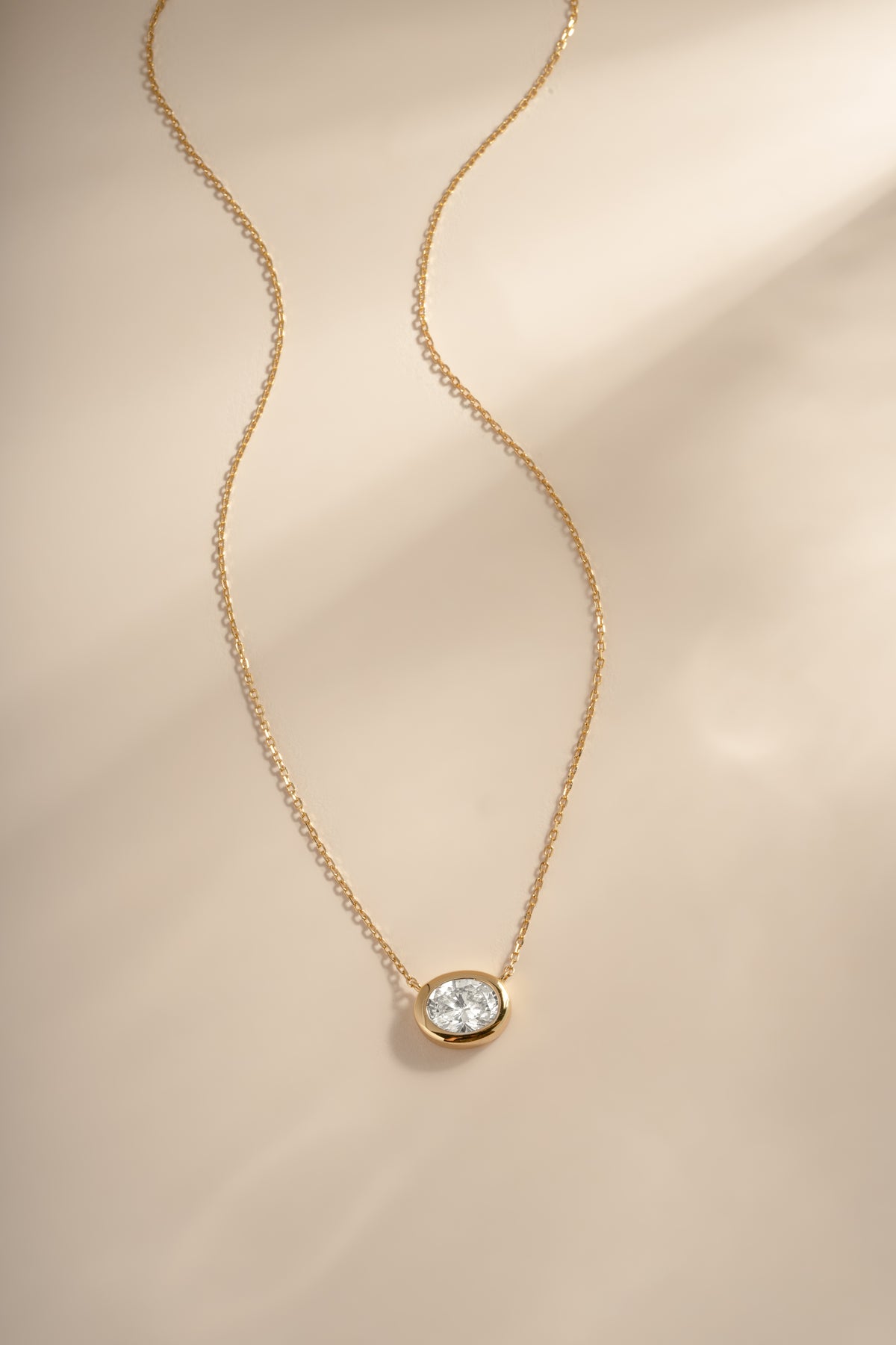Gold east-west oval bezel pendant necklace arranged on a cream background.