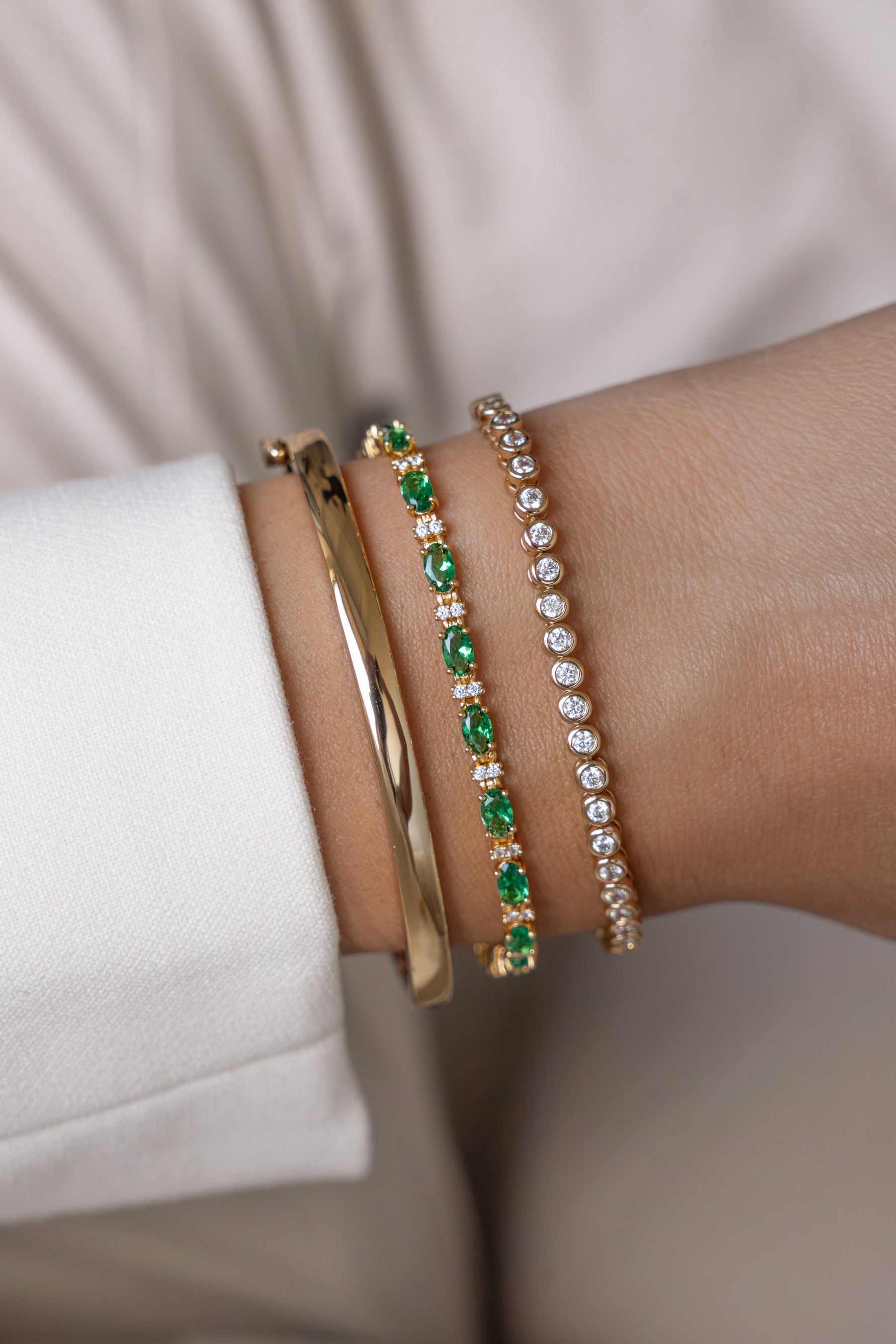 Three stacked bracelets worn on a wrist featuring a gold bangle, a green and white stones tennis bracelet and a gold bezel bracelet.