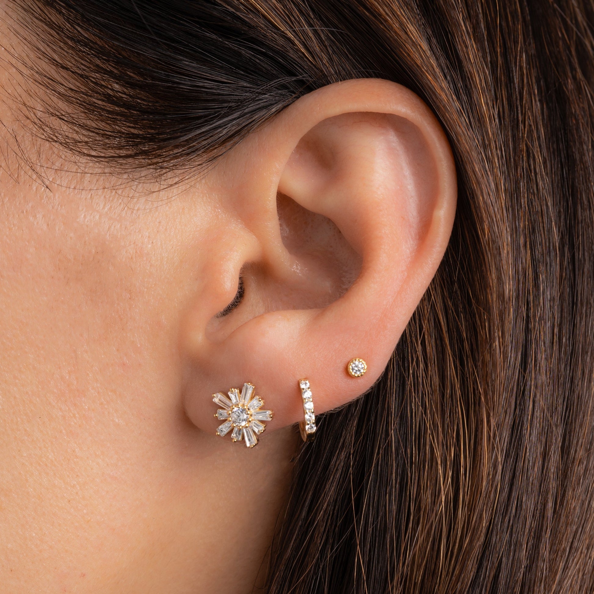 an upclose picture of a model's ear wearing a baguette diamond stud earring, a pave huggie hoop earrings, and a small diamond stud