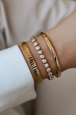 Gold fluted bangle stacked with a round bezel tennis bracelet and a slim smooth bangle on a wrist.
