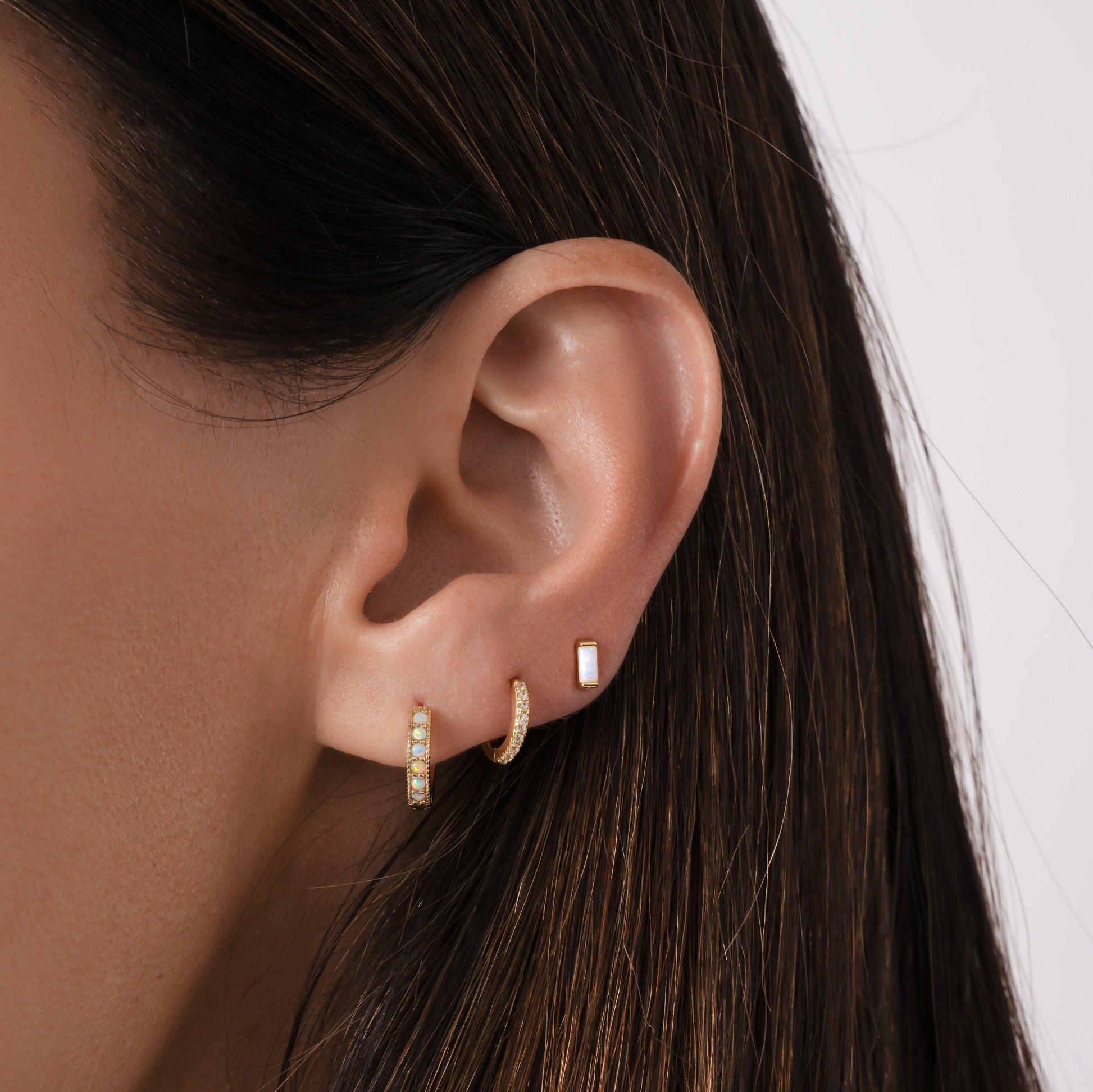 Close up of model's ears with three aesthetic earrings. The first piercing has an opal huggie earring, the second hole has a pave cz baby hoop earring, and the third piercing has a tiny opal stud earring.