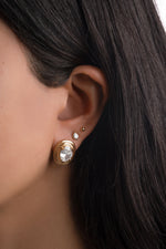 Gold oval bezel statement stud on a model's ear, styled with a pear-shaped stud and a small ball stud.