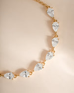 A gold pear tennis bracelet featuring a series of pear-cut clear gemstones in prong settings, displayed on a soft beige background.