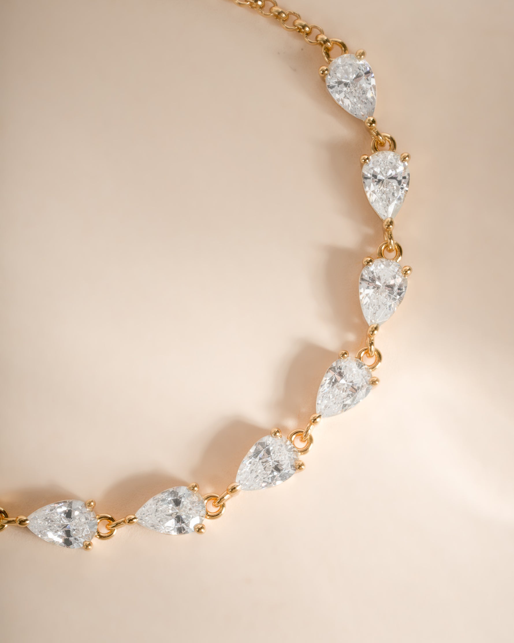 A gold pear tennis bracelet featuring a series of pear-cut clear gemstones in prong settings, displayed on a soft beige background.