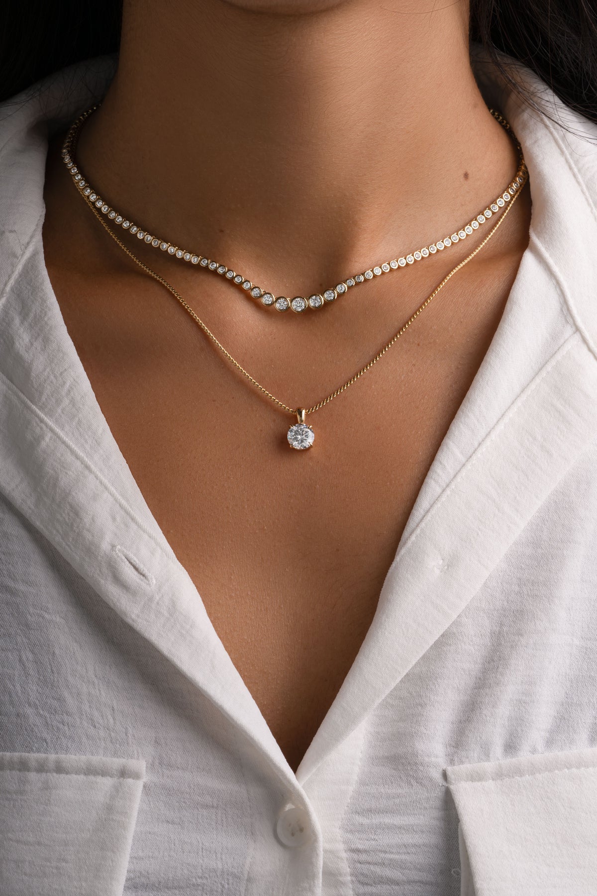 Gold round solitaire pendant layered with a graduated bezel tennis necklace on a model.