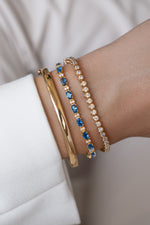 Styled bracelet stack featuring a gold bangle, sapphire tennis bracelet, and bezel bracelet on a wrist.