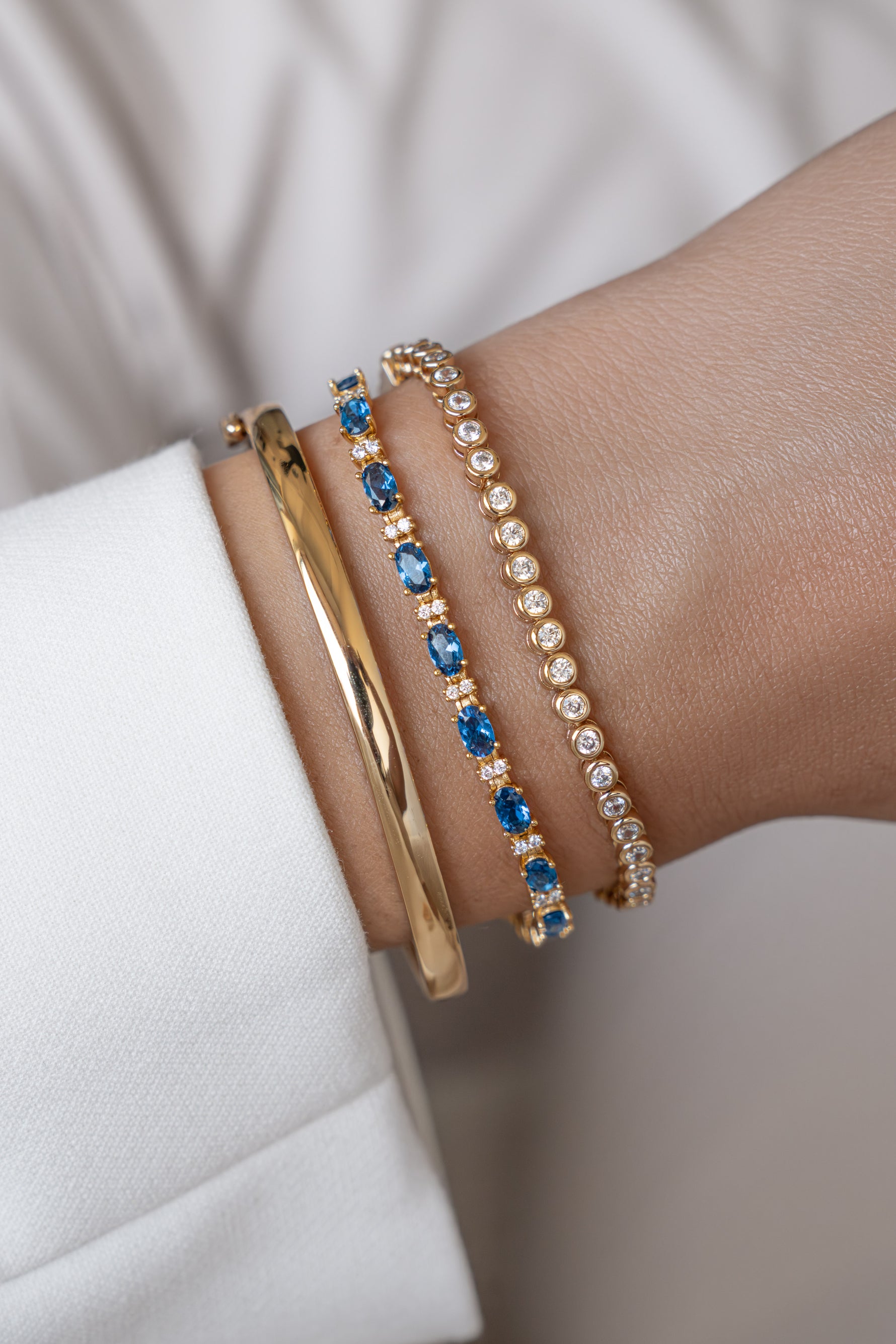 Styled bracelet stack featuring a gold bangle, sapphire tennis bracelet, and bezel bracelet on a wrist.