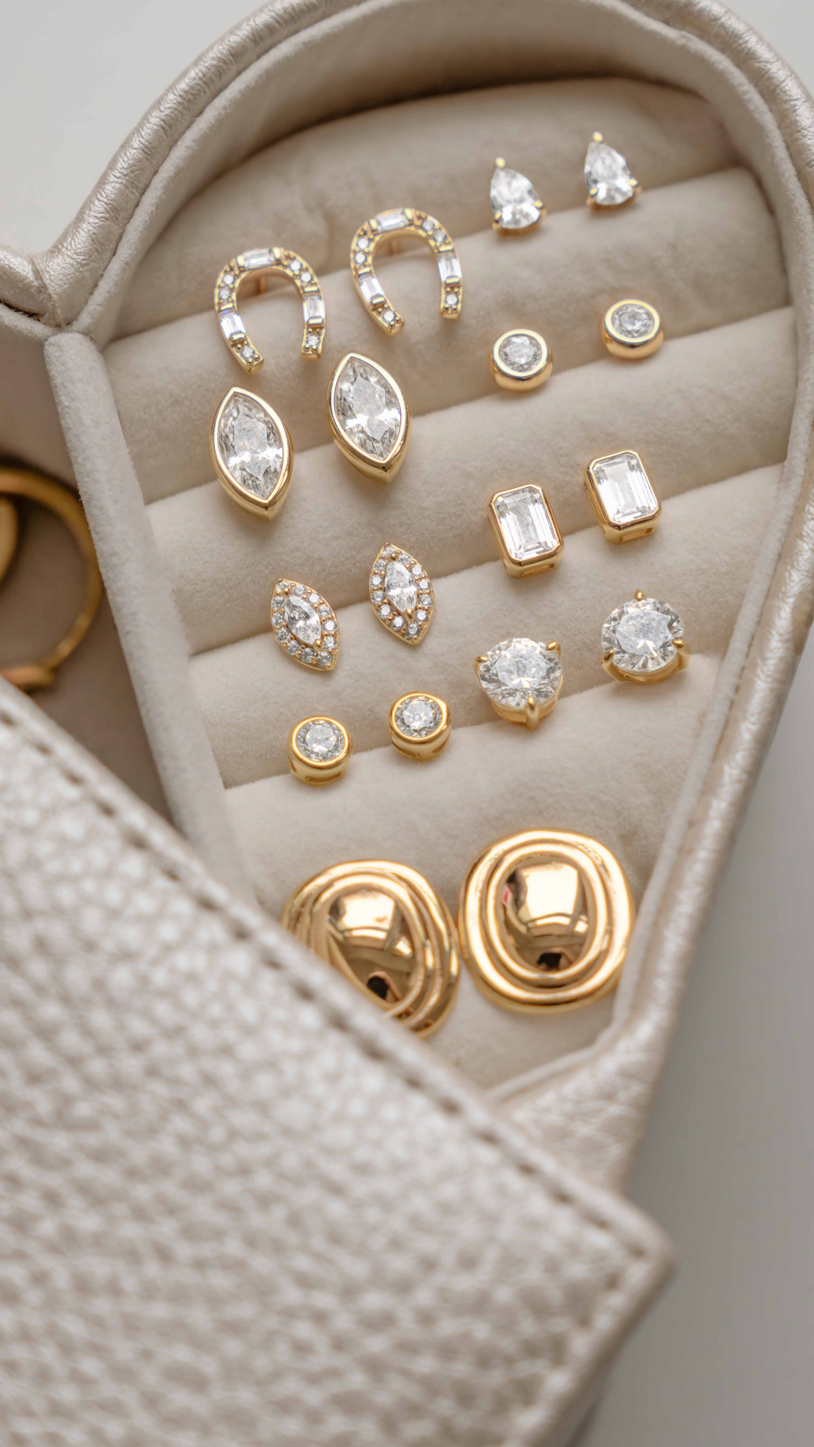 A close-up of a jewelry organizer tray holding multiple gold-tone stud earrings with clear gemstones in different shapes, including marquise, round, rectangular, and halo designs.