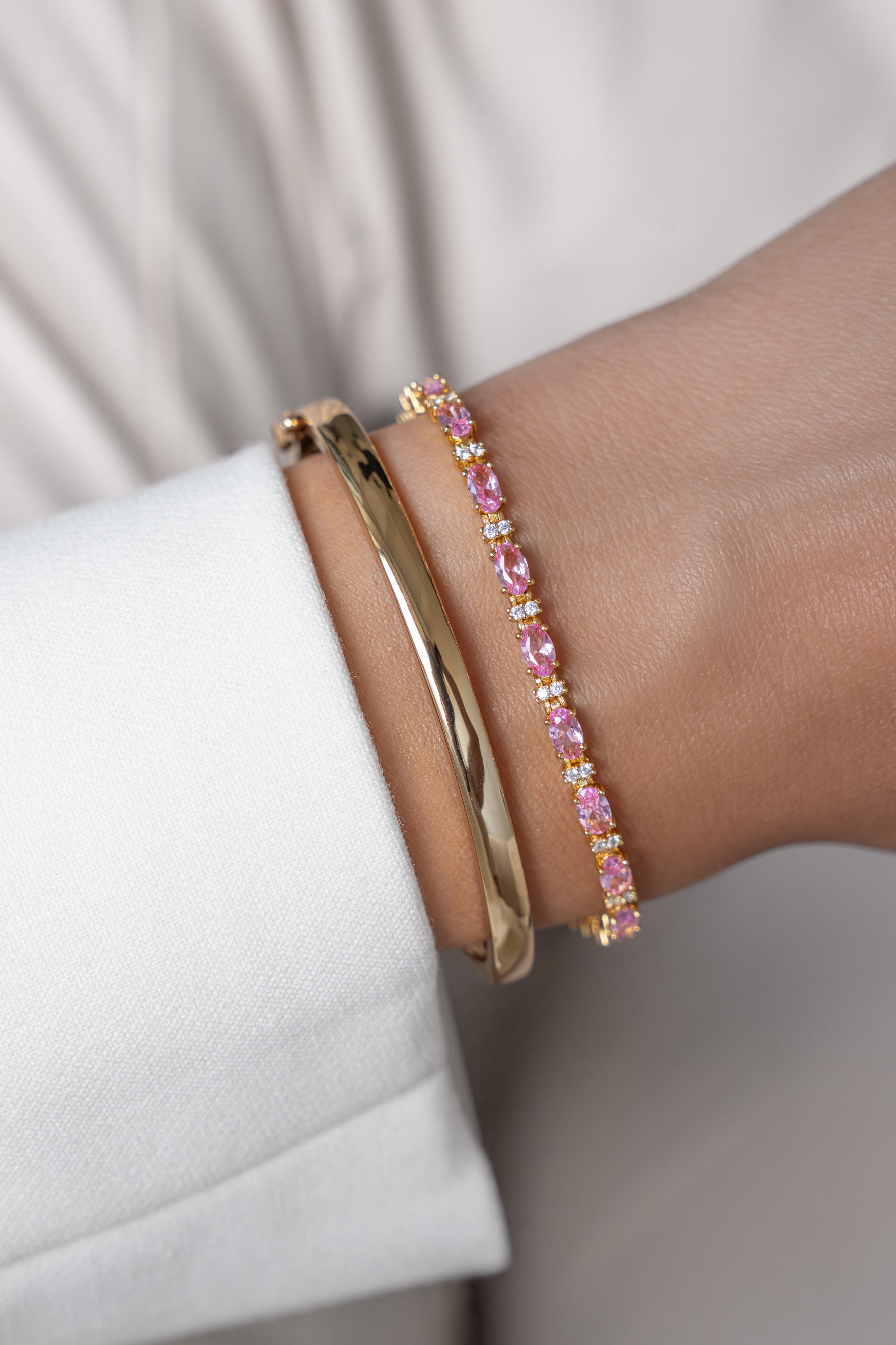 A close-up of a gold bangle and a gold oval pink and white round stones bracelet styled on a wrist.