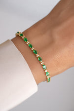 A gold tennis bracelet with green emerald-cut gemstones and small clear accent stones, worn on a wrist. 