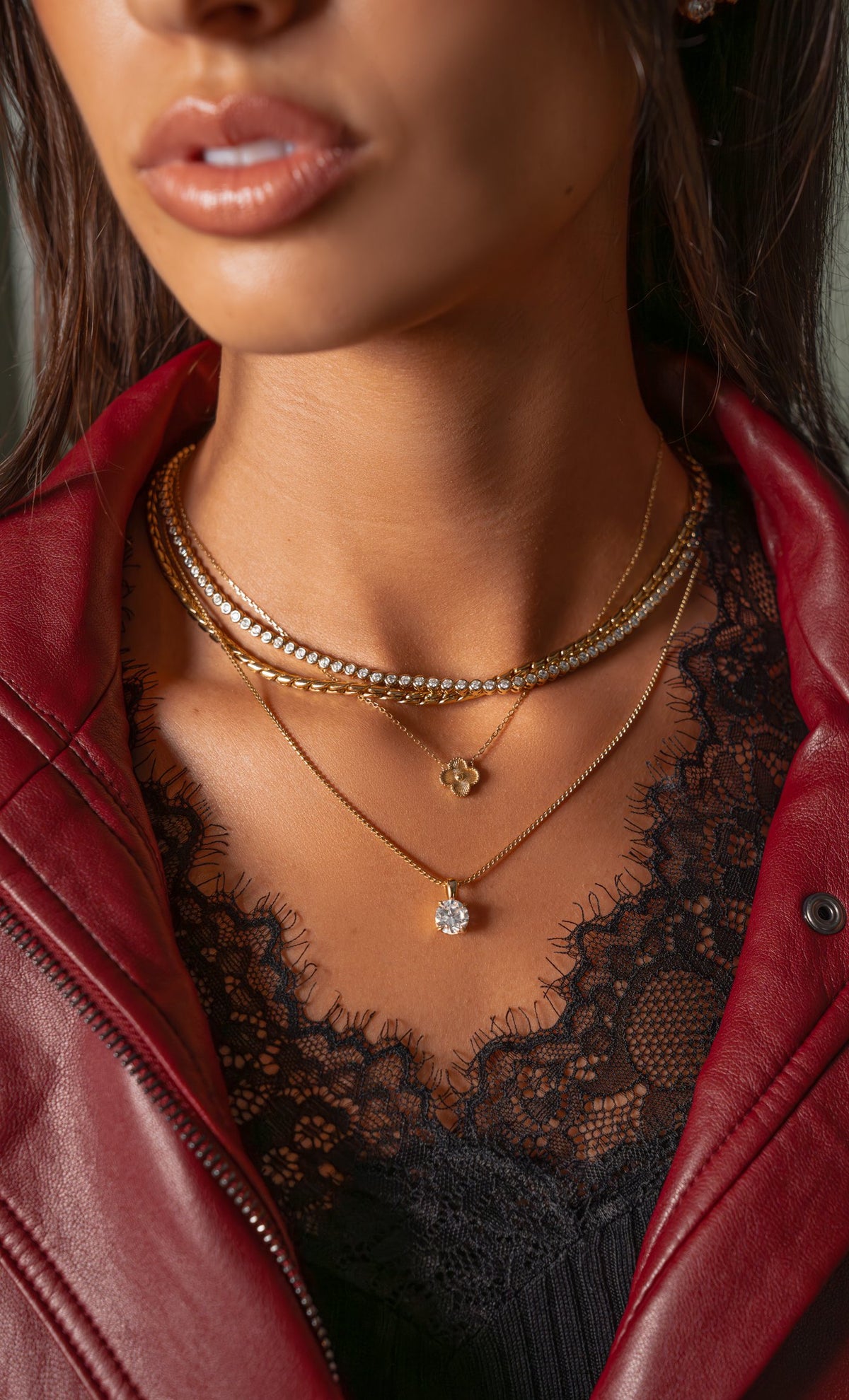 Layered gold necklace stack with bezel tennis necklace, clover and solitaire pendants, worn with a red leather jacket