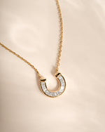 A gold horseshoe pendant necklace adorned with clear baguette-cut stones, displayed on a soft beige fabric background.