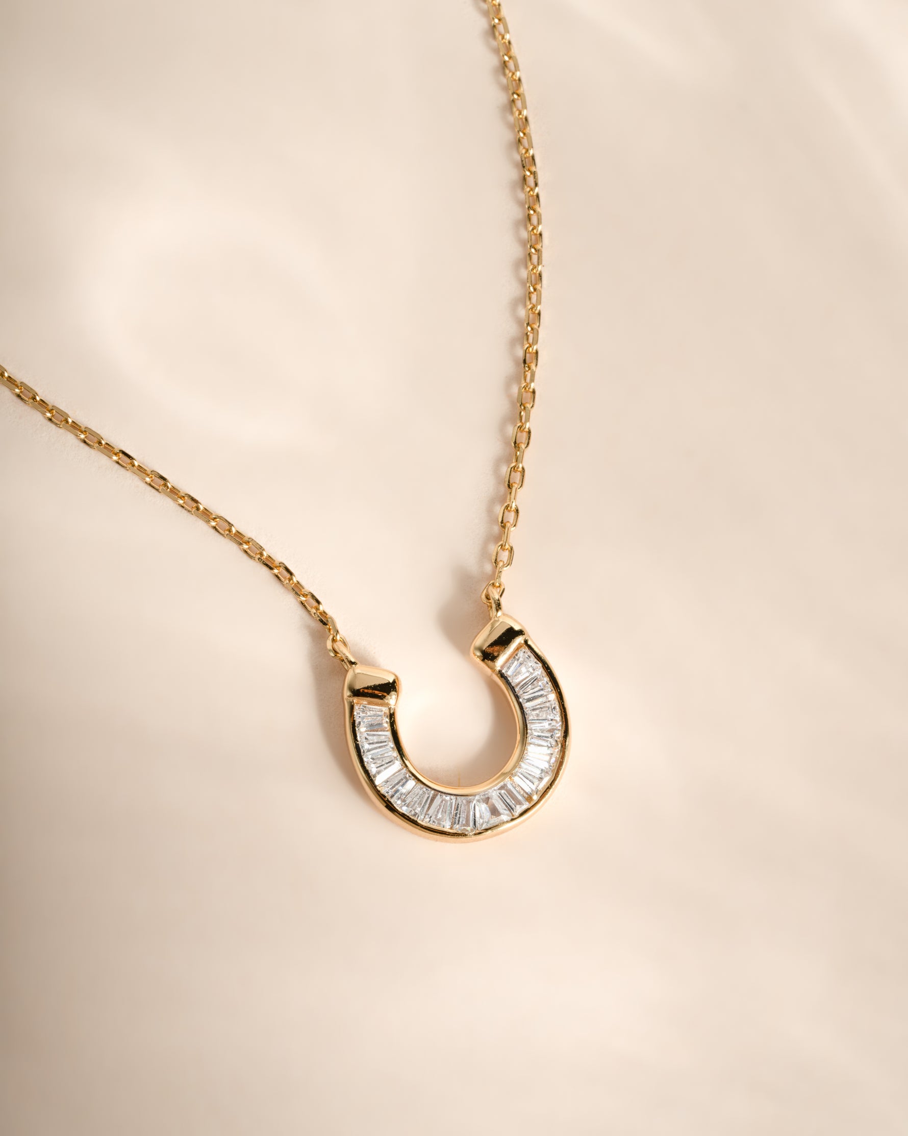 A gold horseshoe pendant necklace adorned with clear baguette-cut stones, displayed on a soft beige fabric background.
