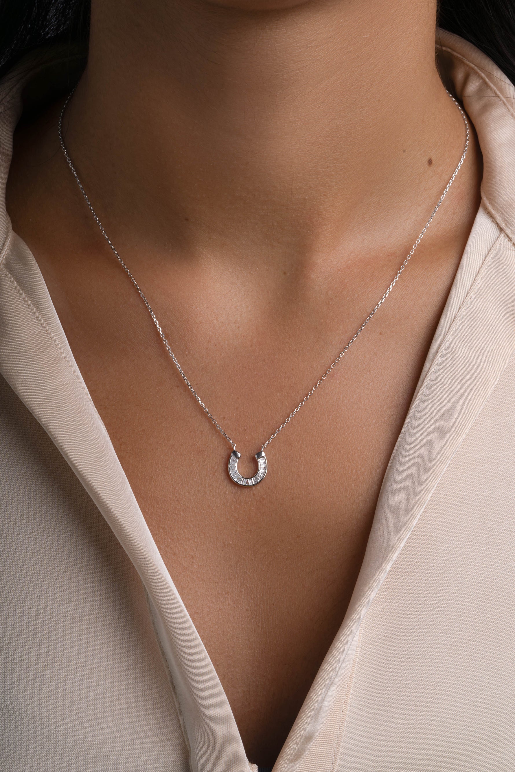 A delicate silver horseshoe pendant necklace worn by a model in a light beige blouse.
