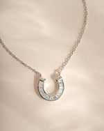 A silver horseshoe pendant necklace paved with clear baguette-cut stones, resting on a soft beige satin fabric background.