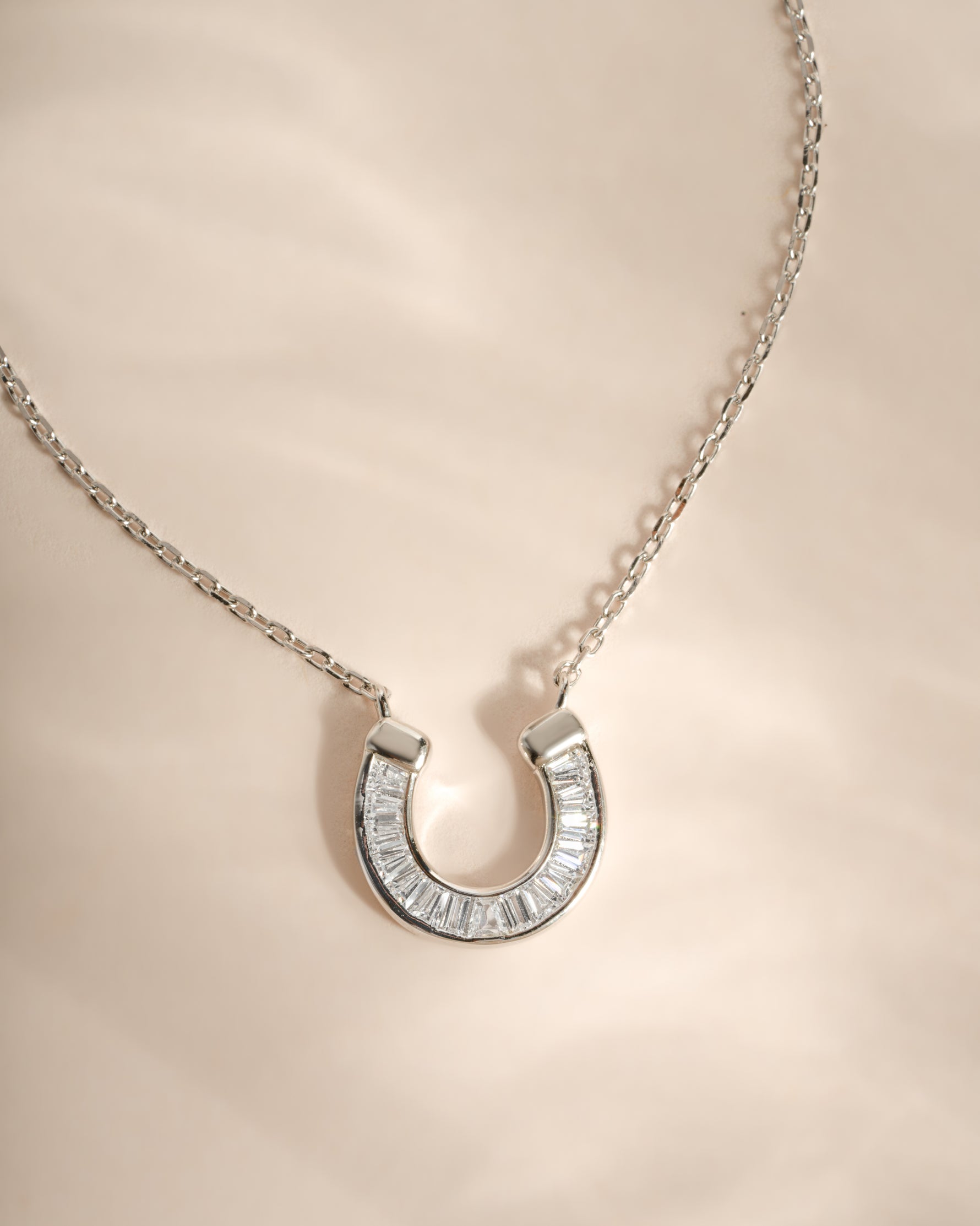 A silver horseshoe pendant necklace paved with clear baguette-cut stones, resting on a soft beige satin fabric background.