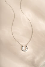 A silver horseshoe pendant necklace with a row of small clear baguette diamond stones, displayed on a delicate silver chain over a soft, light beige satin fabric background.