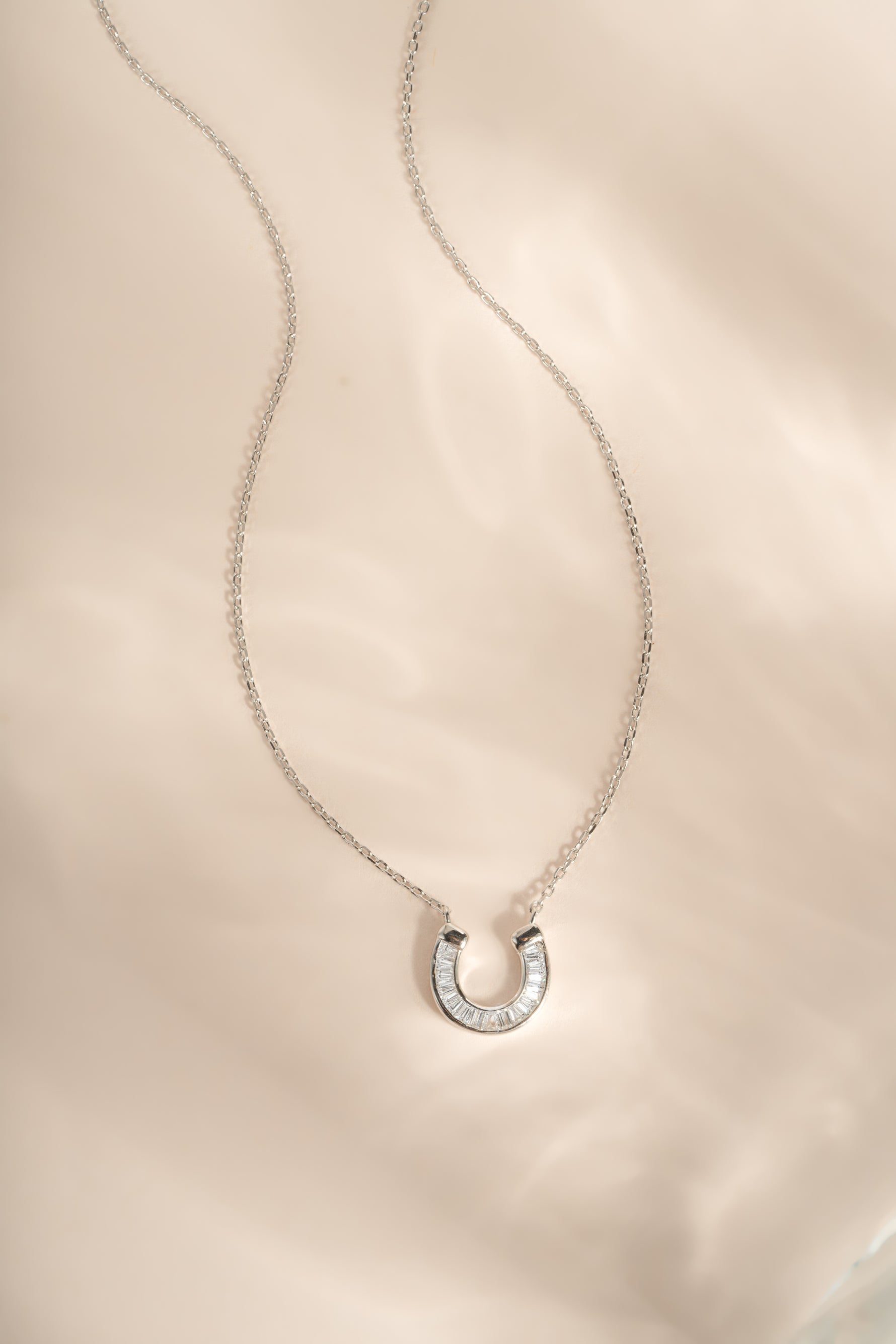 A silver horseshoe pendant necklace with a row of small clear baguette diamond stones, displayed on a delicate silver chain over a soft, light beige satin fabric background.