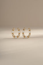 A pair of gold horseshoe shaped stud earrings embellished with clear baguette and round diamonds, displayed on a neutral beige background.