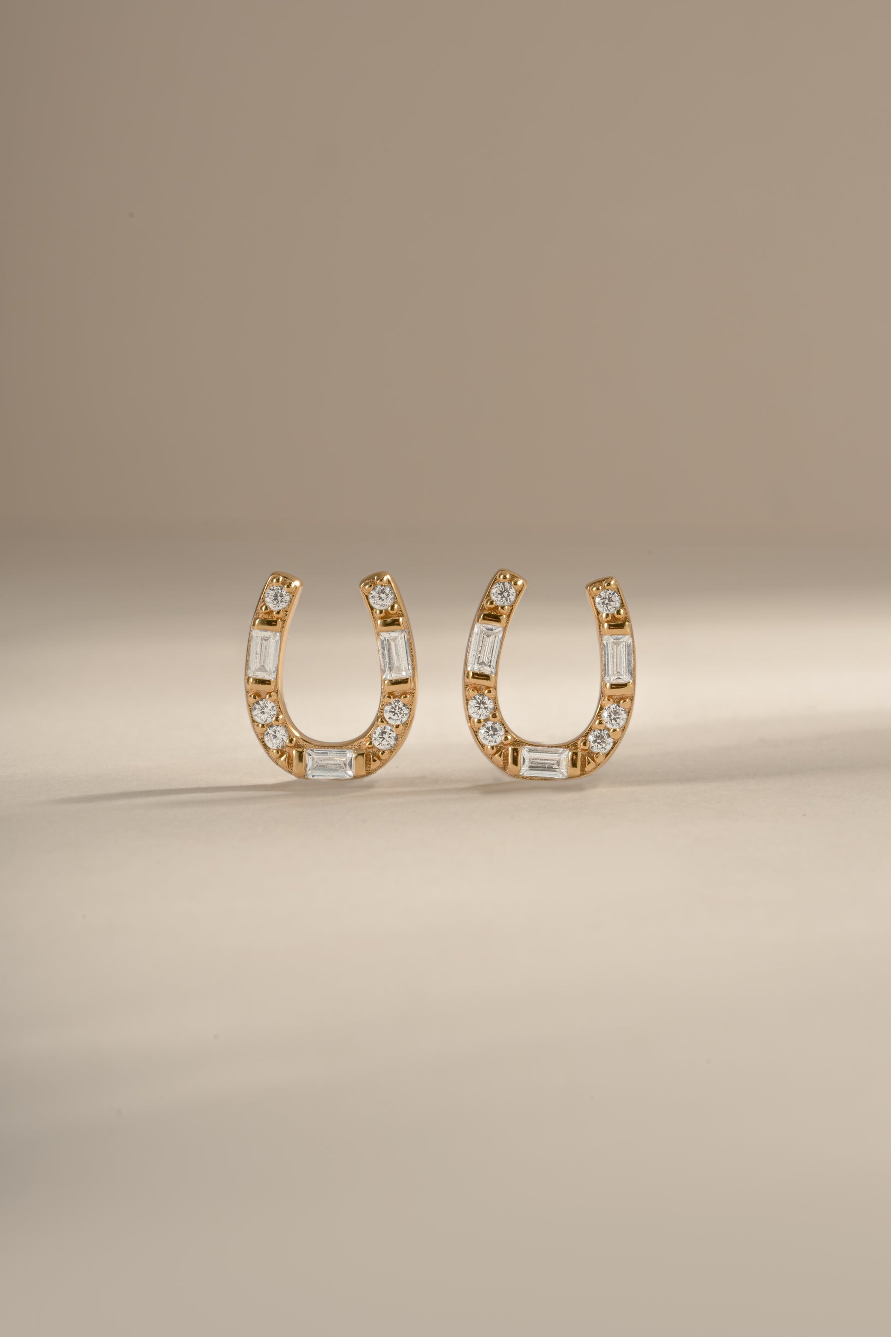 A pair of gold horseshoe shaped stud earrings embellished with clear baguette and round diamonds, displayed on a neutral beige background.